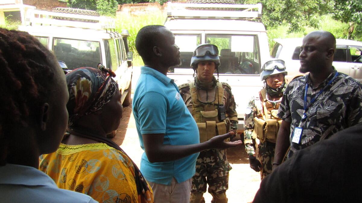 Small group of people standing and talking near parked vehicles, including two individuals in camouflage uniforms and protective gear.