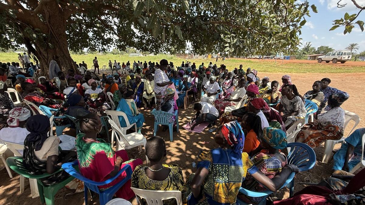 A large group of people sit in a circle under a tree during an outdoor community meeting.