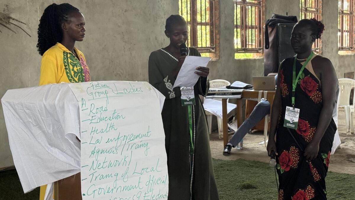 Three individuals standing indoors during a group activity, with one reading from a paper beside a flip chart listing topics such as roads, education, health, and law enforcement.