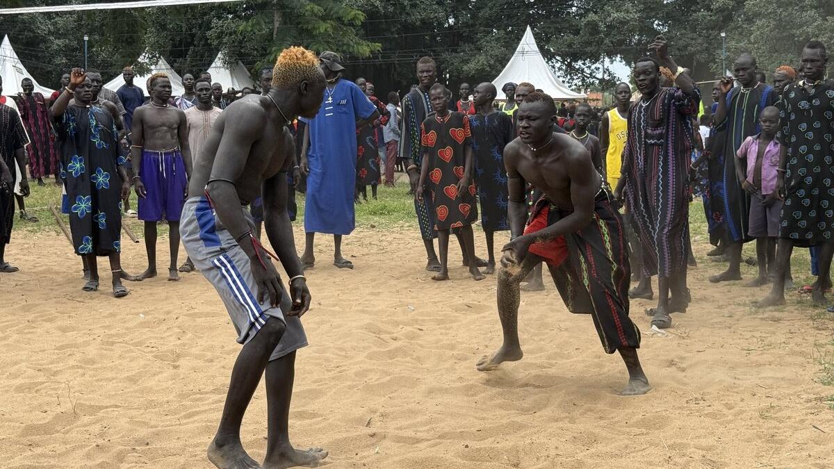 Two individuals engaged in a traditional wrestling match on sandy ground, surrounded by a crowd wearing colorful patterned clothing, with white tents and trees in the background.