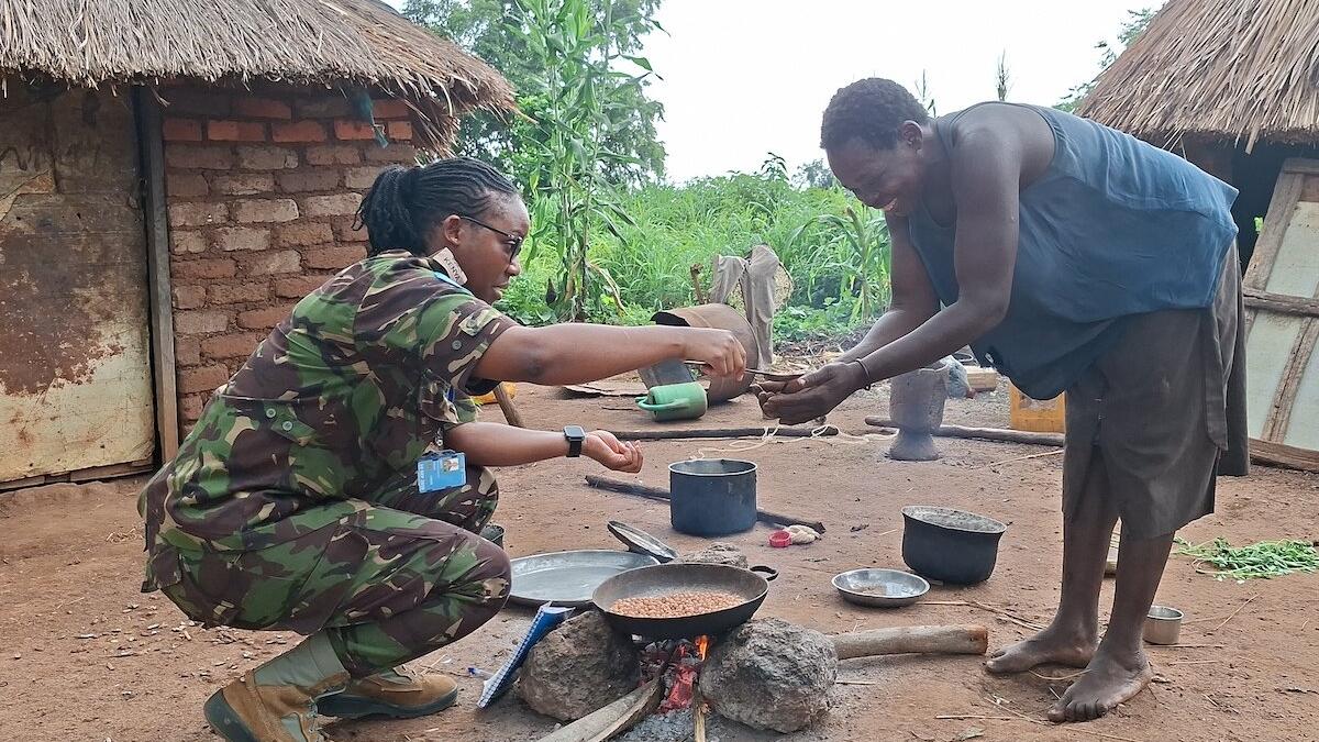 A person in camouflage uniform kneels beside an outdoor cooking area, handing something to another person near a fire with pots and a pan in front of traditional huts