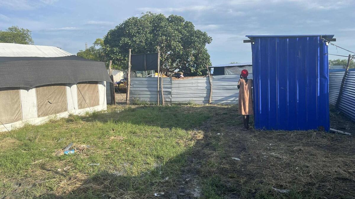 Outdoor area with grass and mud, showing a blue metal structure next to a fenced compound with tents and trees in the background. In the distance, a woman stands wearing an orange dress.