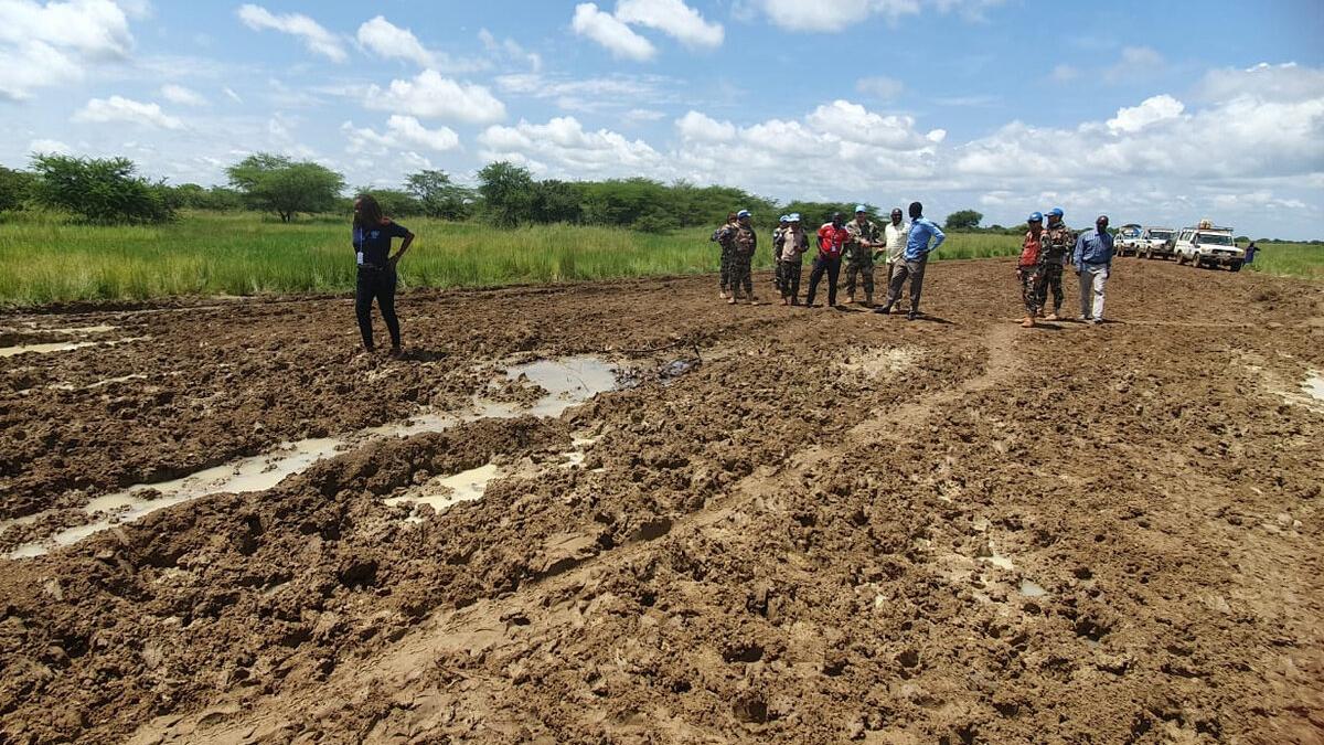 A group of people, including uniformed personnel, stand on a muddy road with puddles, near grassy fields and parked vehicles under a partly cloudy sky.