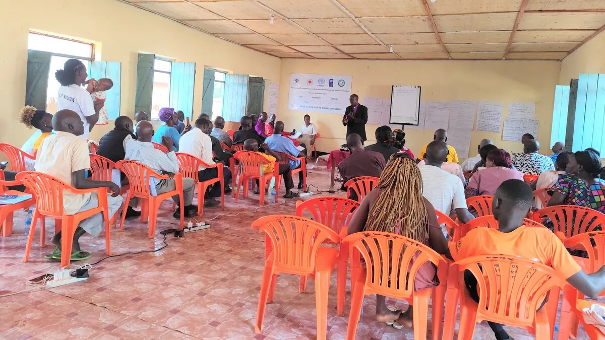 A community meeting in a room with orange plastic chairs arranged in rows, participants seated and listening to a speaker at the front near posters and a banner on the wall.