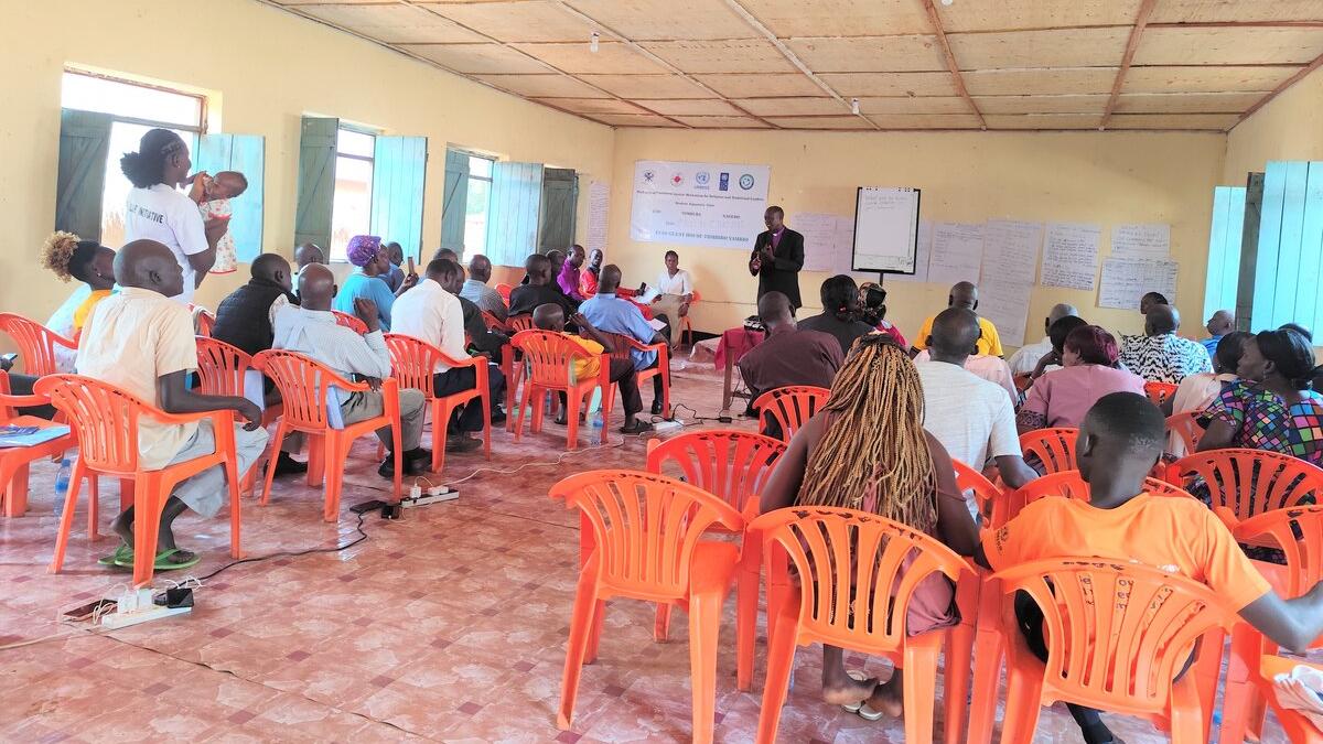 A community meeting in a room with orange plastic chairs arranged in rows, participants seated and listening to a speaker at the front near posters and a banner on the wall.