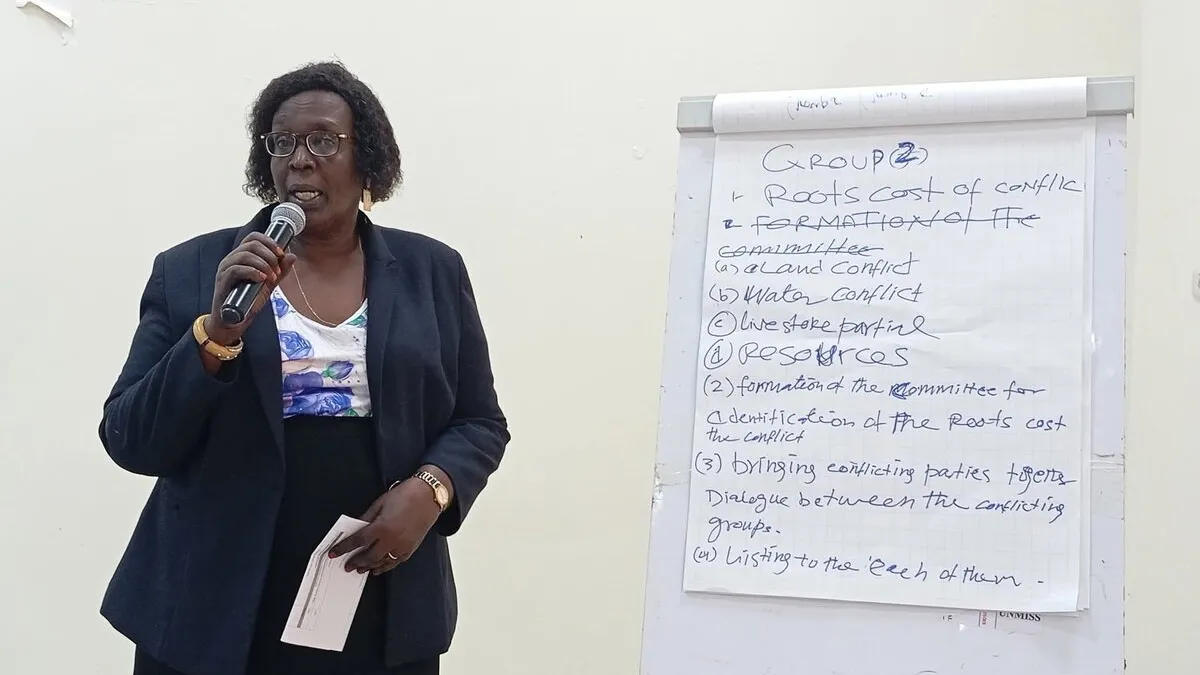 Person holding a microphone and standing beside a flip chart with handwritten notes on conflict causes and committee formation.