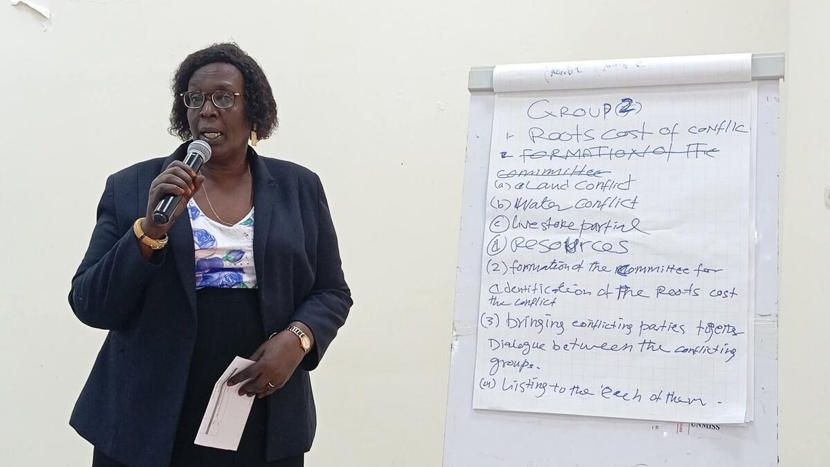 Person holding a microphone and standing beside a flip chart with handwritten notes on conflict causes and committee formation.