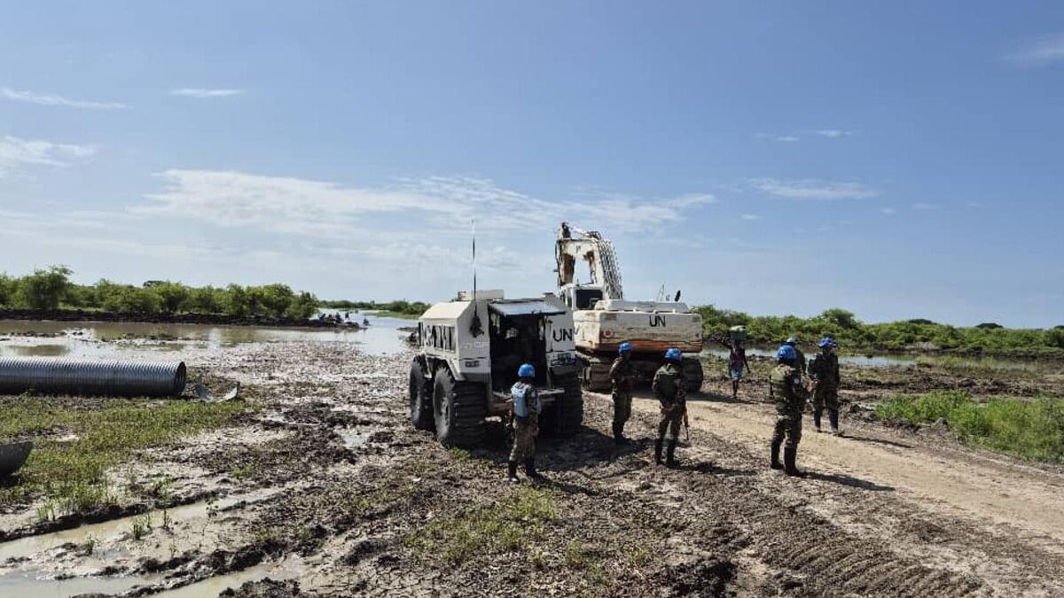 UN peacekeepers in blue helmets stand on a muddy road near heavy UN machinery and pipes, with water and greenery in the background.