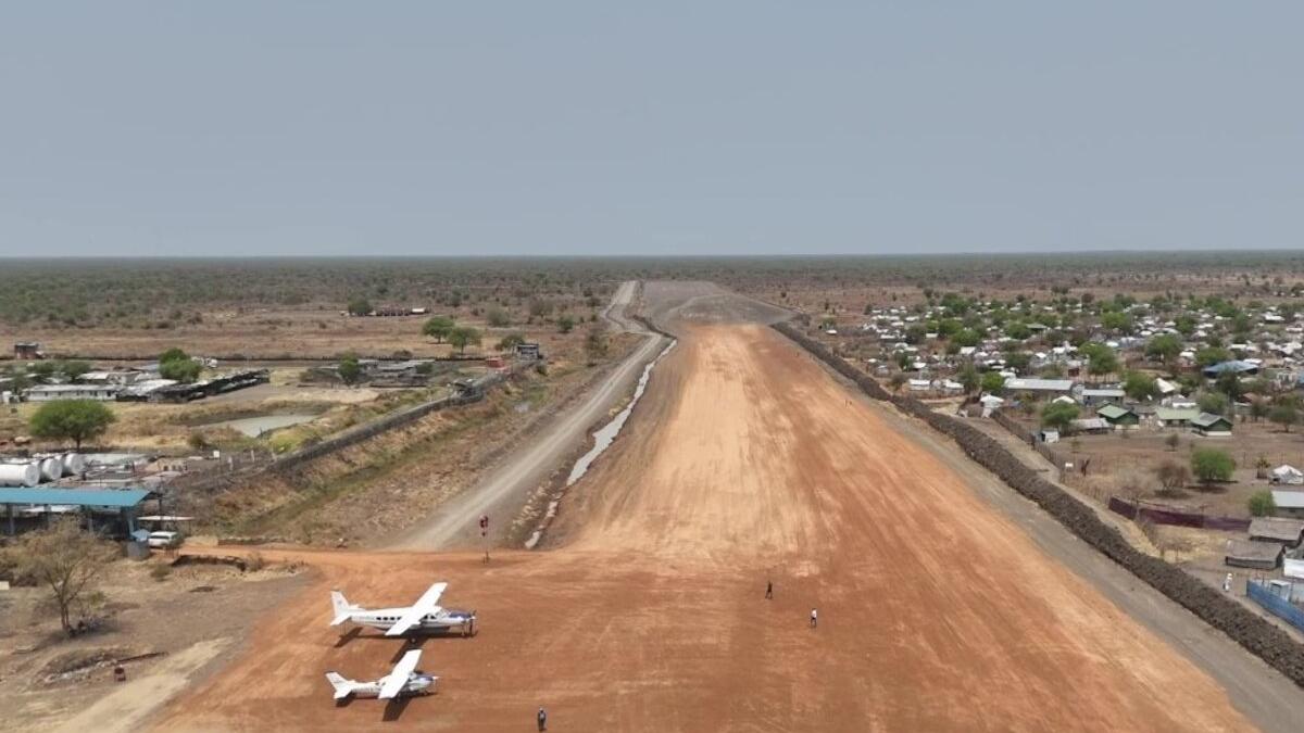 A dirt airstrip with two small white aircraft parked at one end, surrounded by sparse vegetation and scattered buildings under a clear sky in a remote area.