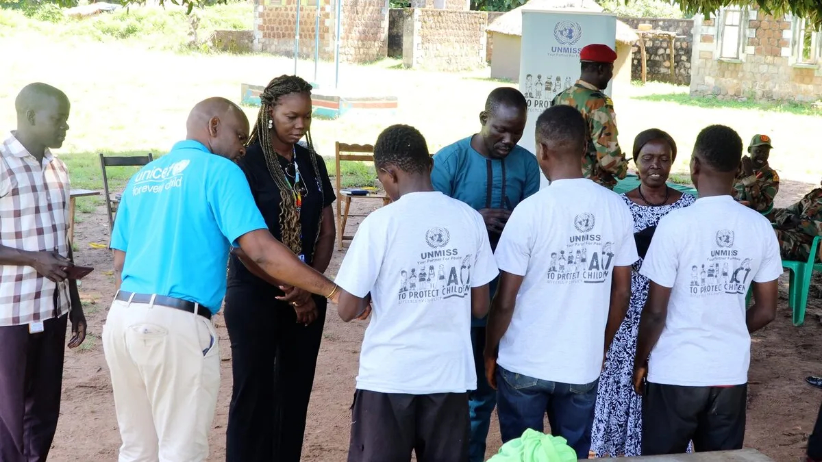 Three children stand under mango trees with a few belongings as they are released from South Sudanese armed forces and begin a new life. Group of people standing outdoors in a circle, including individuals wearing UNMISS-branded shirts and a person in a UNICEF shirt, with soldiers and chairs in the background.