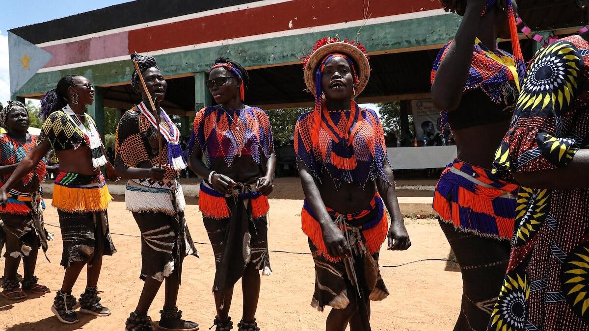 Group of people wearing colorful traditional attire with beadwork and skirts, performing a cultural dance outdoors in front of a painted structure.
