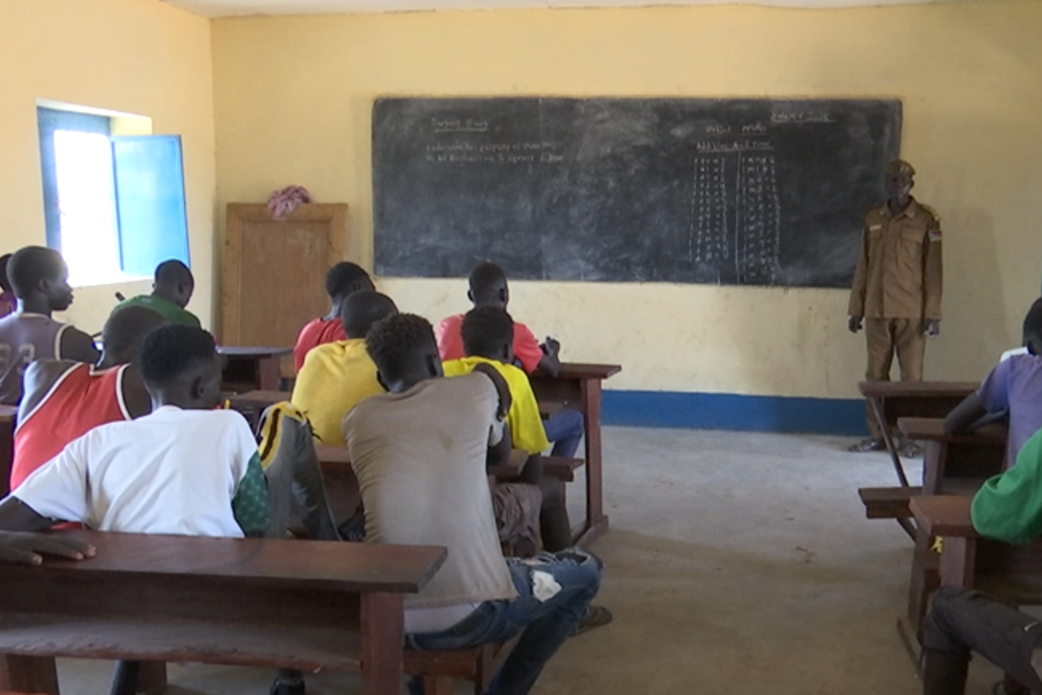 At the prison in Kuajok, the vocational trainings initiated by UNMISS previously have an impact by offering inmates to develop skills in tailoring, farming and accounting. Classroom scene with students seated at wooden desks facing a chalkboard, where mathematical calculations and notes are written. An instructor stands near the board in front of the class.