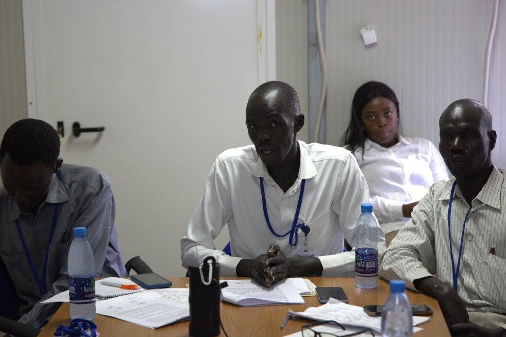 Several people seated at a table indoors during a meeting, with notebooks, papers, and water bottles spread across the table.