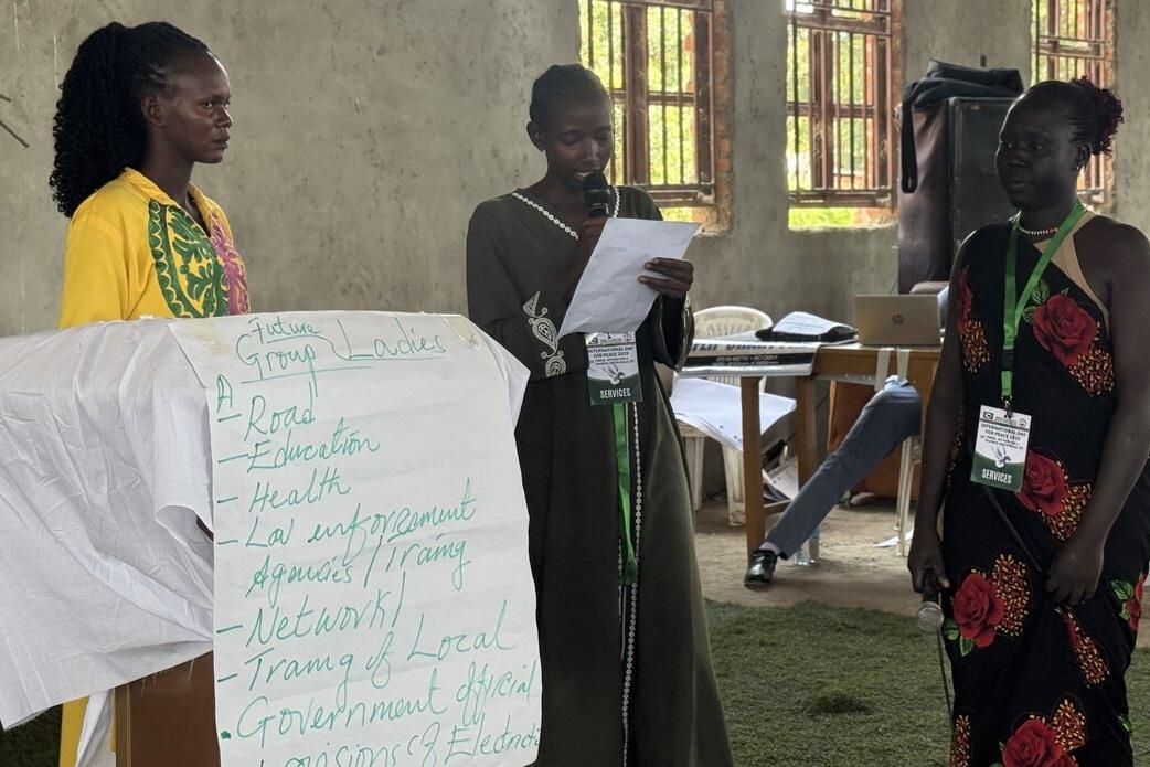 Three individuals standing indoors during a group activity, with one reading from a paper beside a flip chart listing topics such as roads, education, health, and law enforcement.