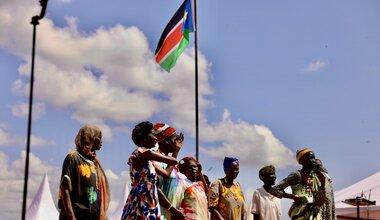 Group of individuals outdoors singing near a flagpole with the South Sudan flag, under a partly cloudy sky.