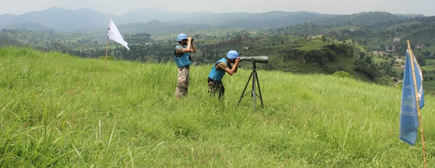 Two UN peacekeepers wearing blue helmets and vests observe terrain from a grassy hilltop using binoculars and a tripod-mounted scope.